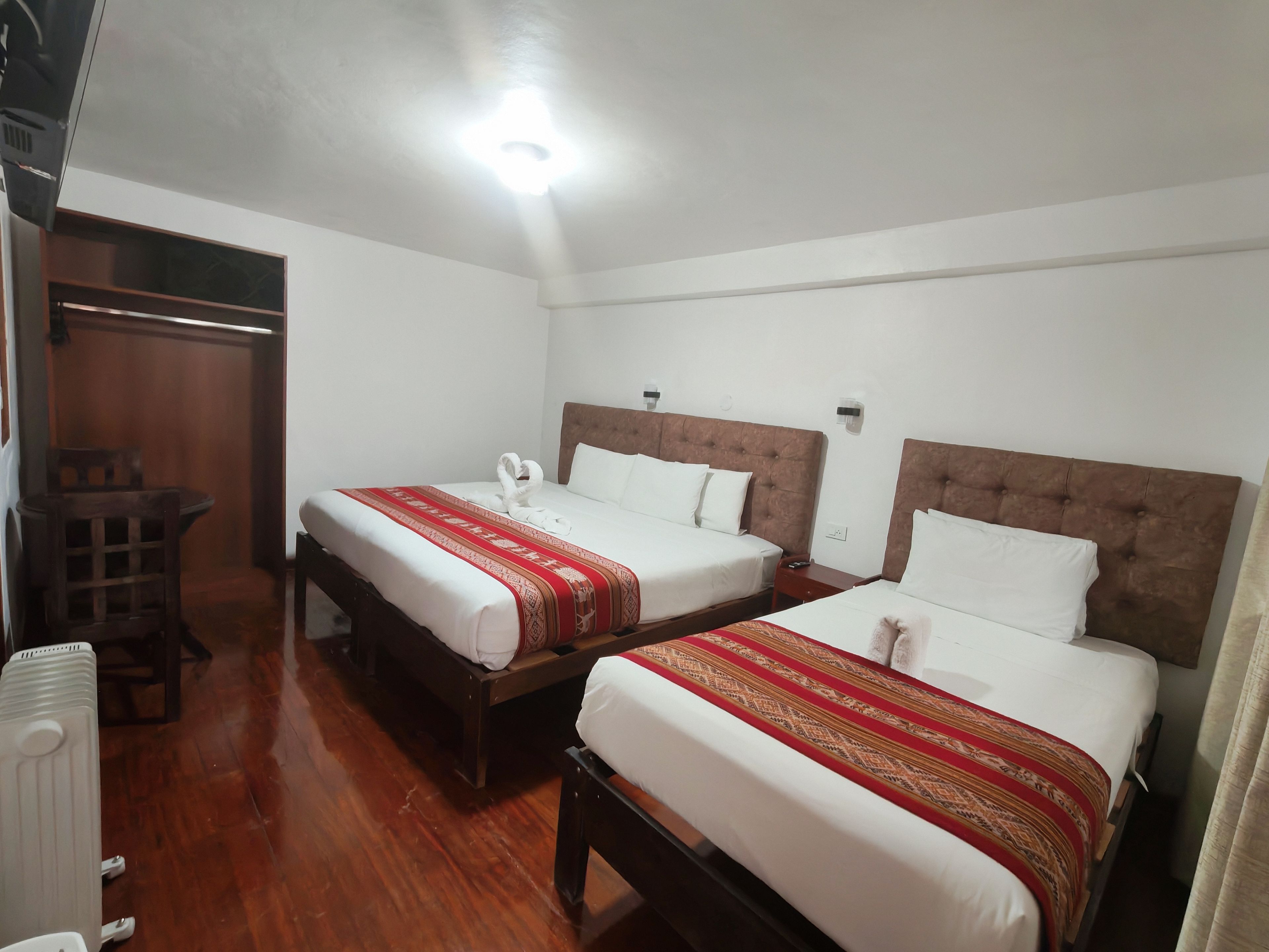Habitación de hotel con dos camas, cabeceros de madera, ropa de cama blanca, colcha estampada roja, una mesita con una silla y un radiador.