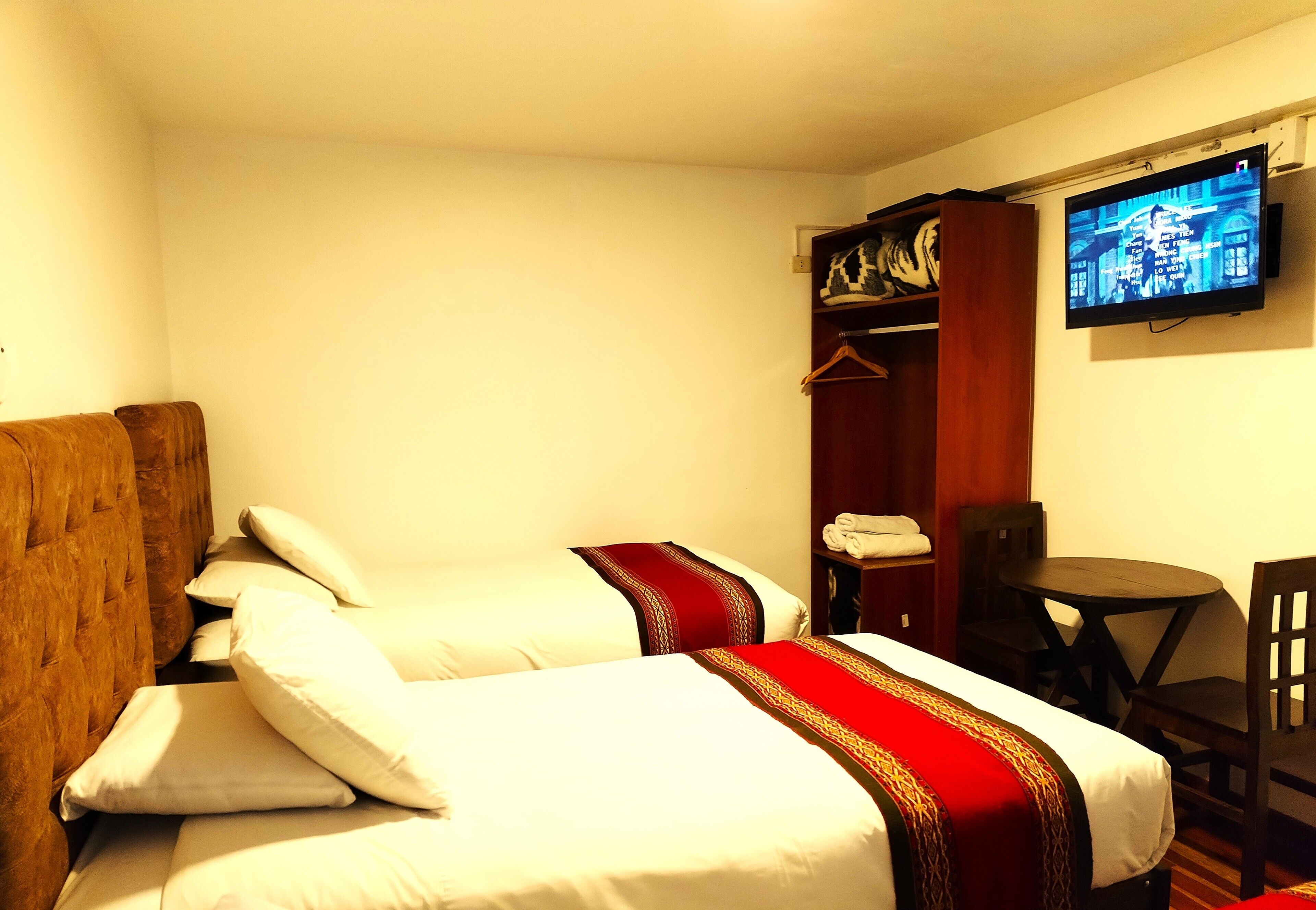 Habitación de hotel con dos camas, cabecera de madera, televisor montado en la pared, una mesita y una silla.