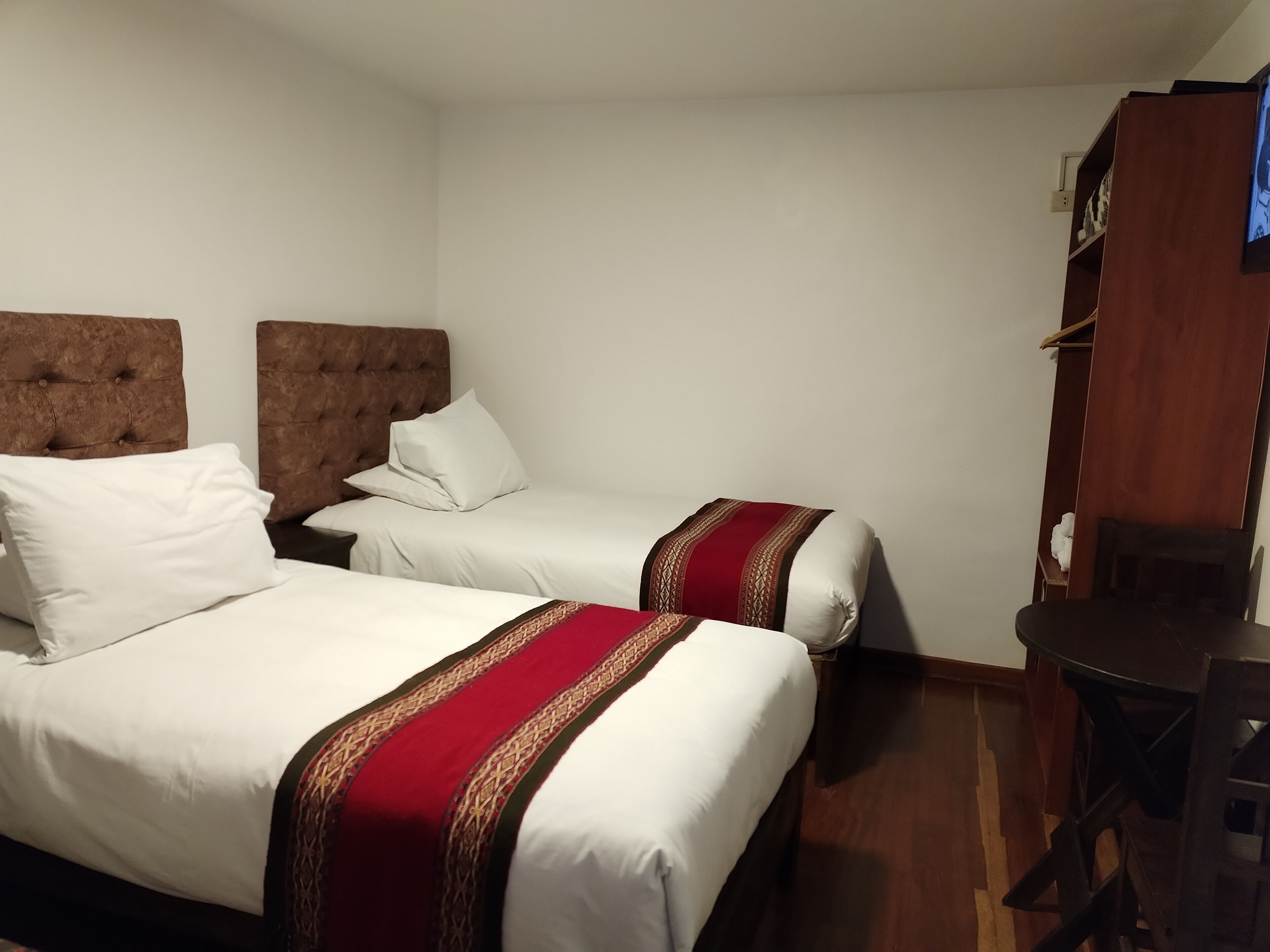 A hotel room with two beds, each with a headboard, white bedding, and a red and brown decorative trim. There is a wooden desk and a television on the wall.