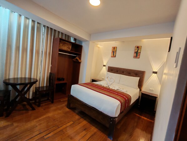A hotel room with a wooden floor, a bed with white bedding and a red patterned blanket, a small round table, a chair, a wardrobe, and wall-mounted lamps. - Paqarina san Blas (Cusco)