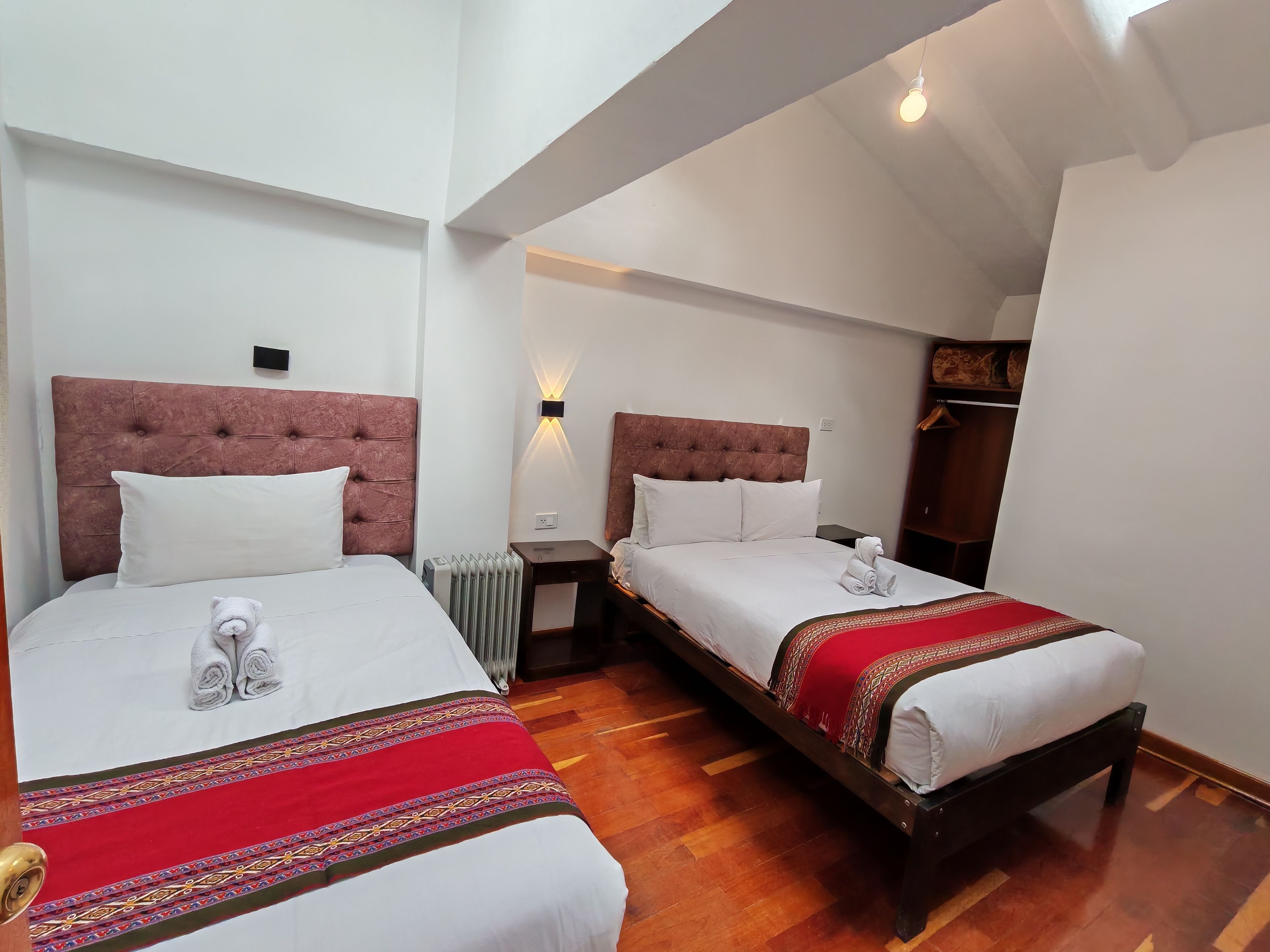 A hotel room with two beds, each with a red and white bedspread, a wooden headboard, and a white pillow. There is a nightstand with a lamp between the beds, and a radiator on the floor.