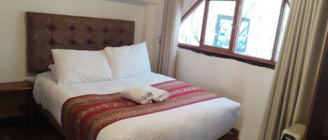 A neatly made bed with white linens and a red patterned blanket, a wooden headboard, and a window with curtains.