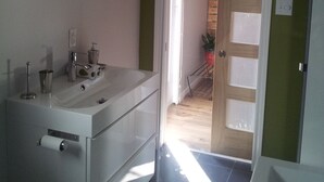 Une salle de bain moderne avec un lavabo blanc, un miroir et un espace douche. Un pot de plante se trouve dans un coin, et une porte donne accès à une autre pièce.