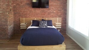 Une chambre avec un mur en briques, un téléviseur à écran plat fixé au-dessus d’un lit en bois, et une fenêtre sur le côté droit.
