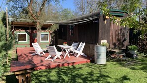 A wooden cabin with a patio area featuring white lounge chairs and a table, surrounded by greenery and trees. - Kouzelná maringotka (Chribska)