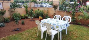 Eine kleine Terrasse im Freien mit Tisch und Stühlen, umgeben von einem Garten mit verschiedenen Pflanzen und Blumen.