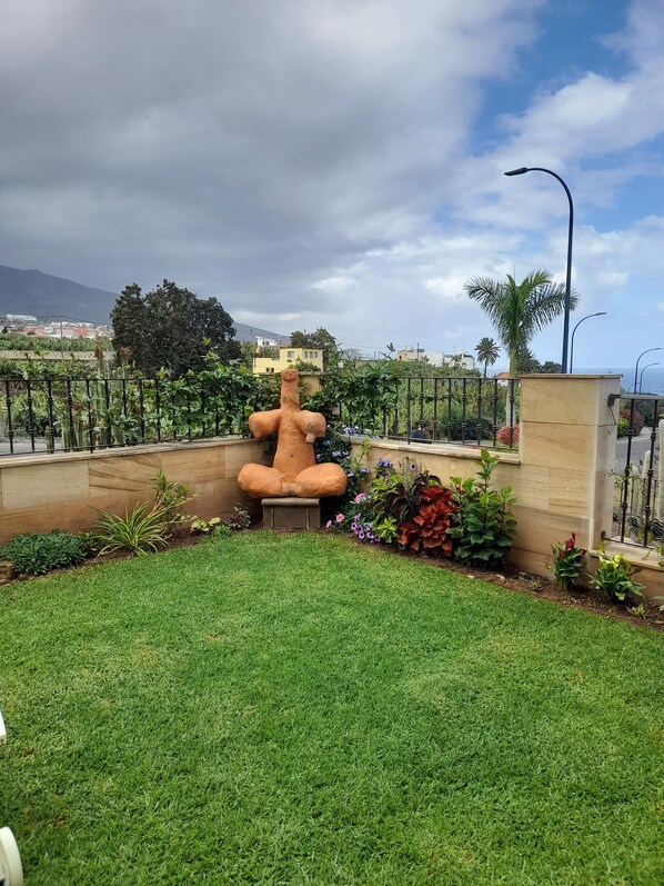 Ein Garten mit einer großen Skulptur, einem gepflegten Rasen und Blick auf Gebäude und Berge im Hintergrund.