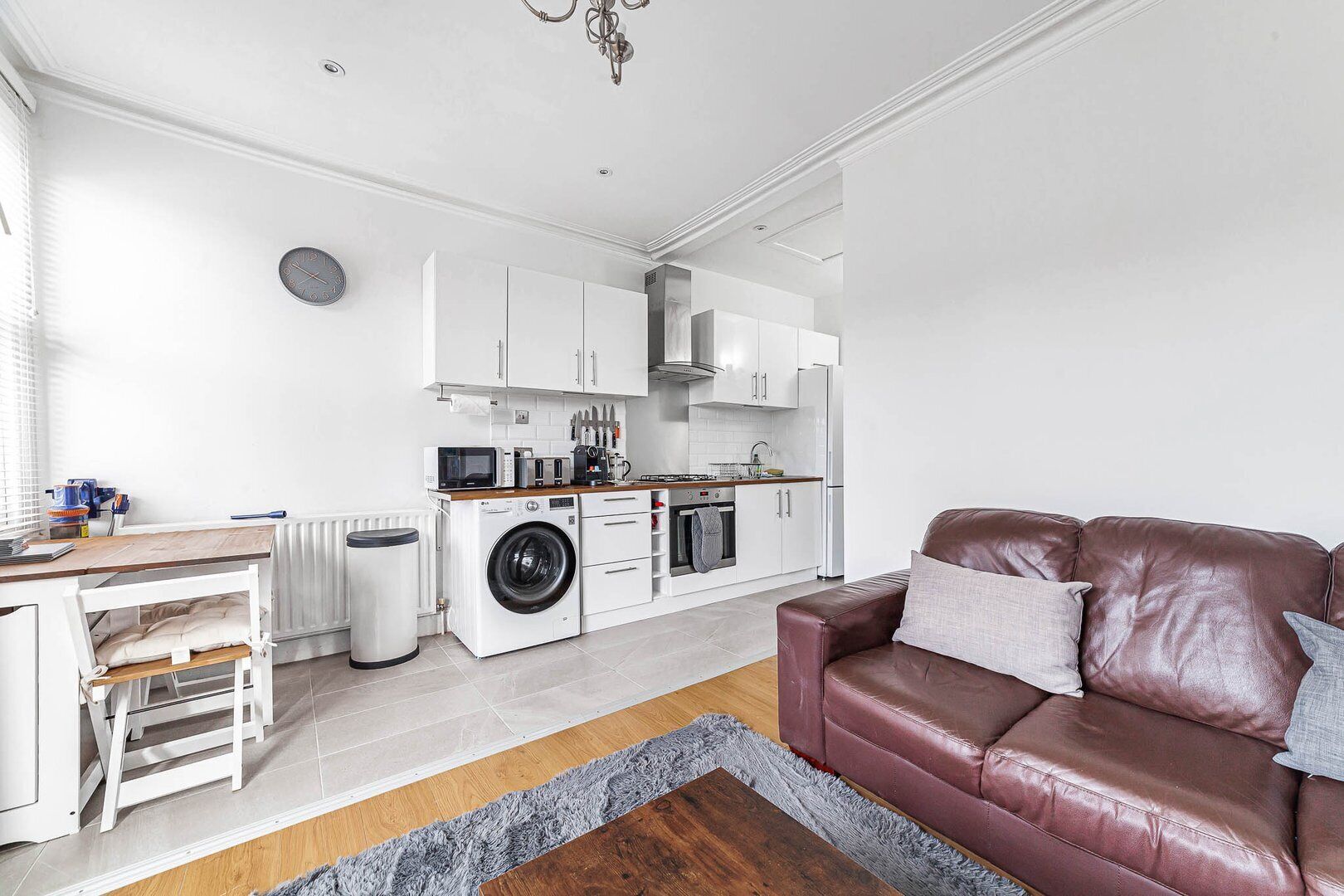 Ett kompakt bostadsutrymme med ett litet kök, en tvättmaskin, ett matbord och en soffa i läder.