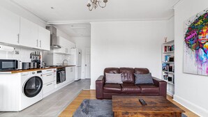 A modern kitchen with white cabinetry, a washing machine, and a dining area with a brown leather couch and a wooden coffee table.