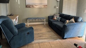 A living room with a blue sofa, a matching armchair, a bench, a flat-screen TV, and a painting of a beach scene.