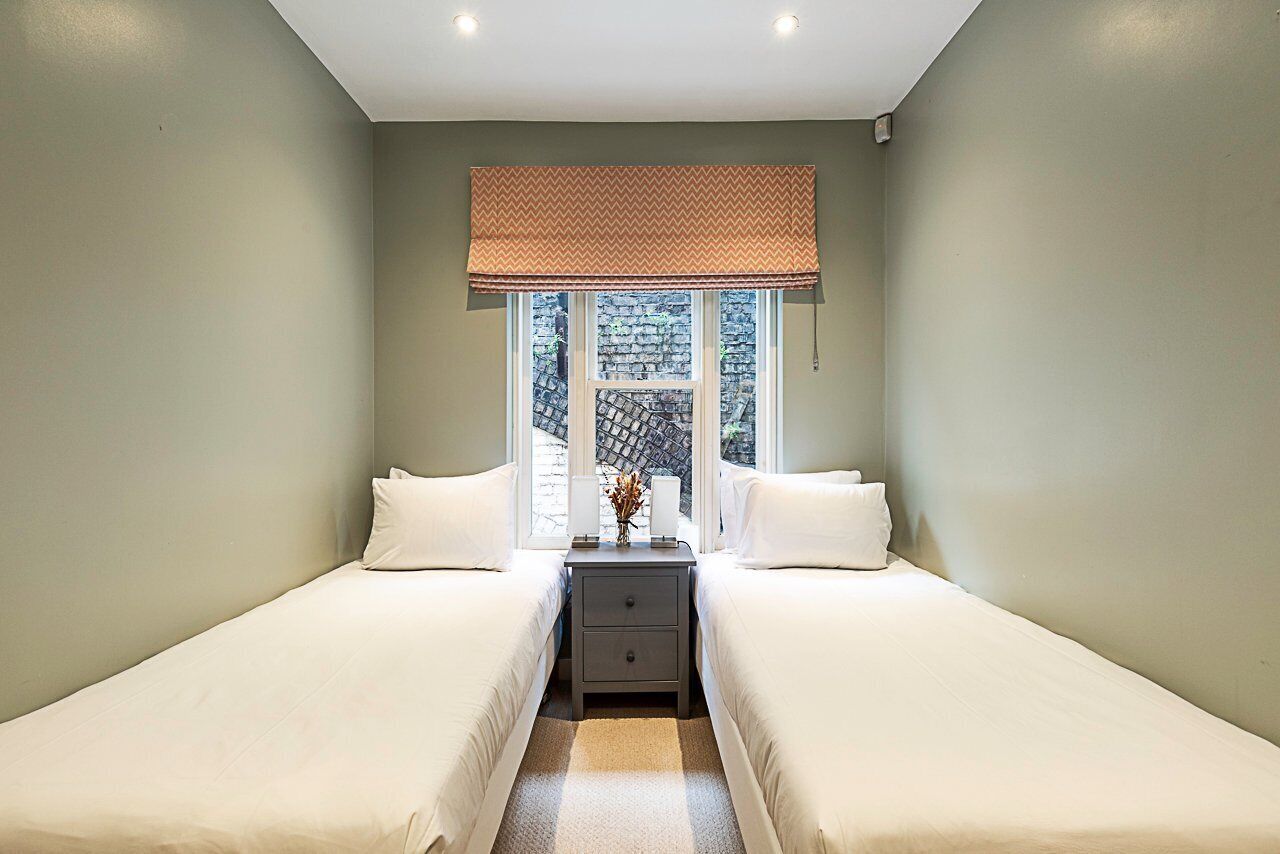 Un dormitorio con dos camas individuales, una mesita de noche con lámpara y una ventana con vista a un muro de ladrillo.