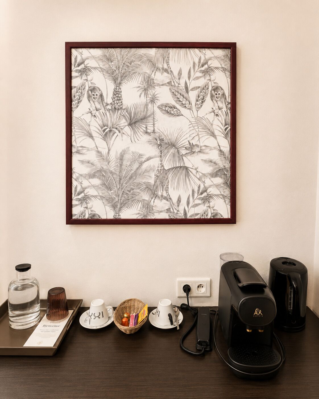 A framed botanical illustration on the wall, a coffee maker, a basket with fruits, a water bottle, and a welcome card on a wooden table.