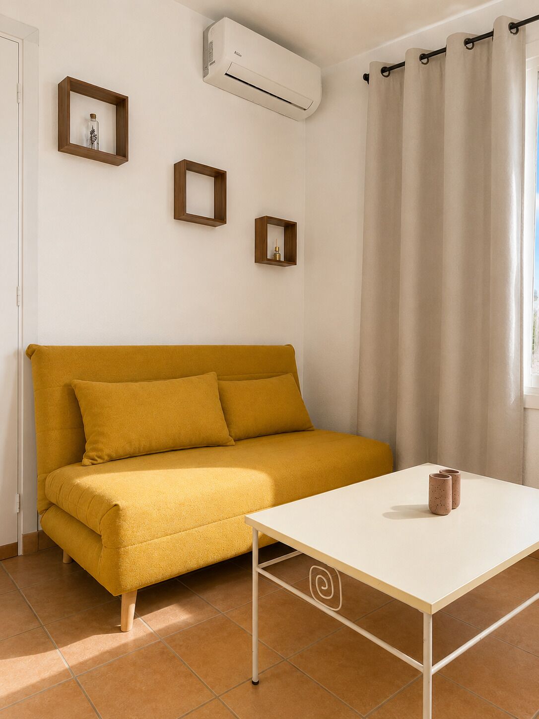 Una habitación con un sofá amarillo, una mesa blanca y una ventana con cortinas.