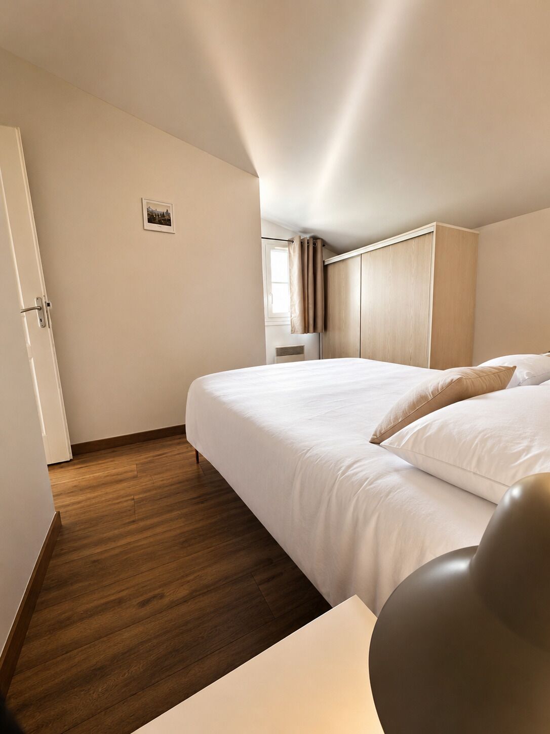 A hotel room with a bed, wooden flooring, a wardrobe, and a small window.