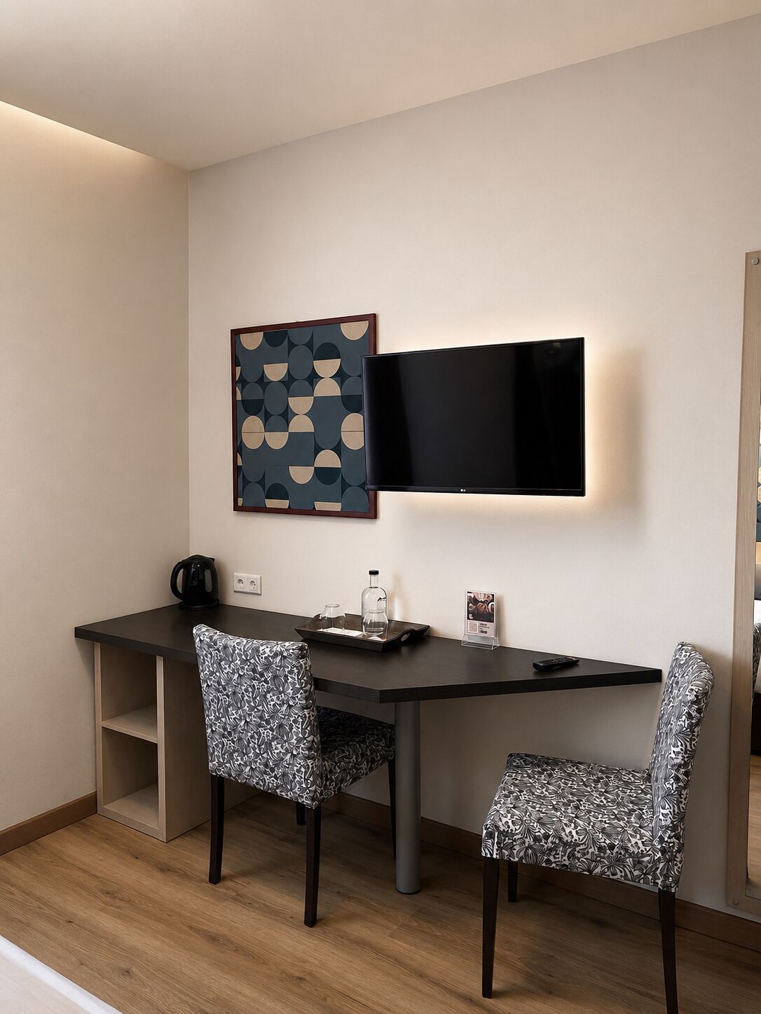 A modern hotel room with a flat-screen TV mounted on the wall, a desk with a tray of amenities, two patterned chairs, and a framed abstract artwork.