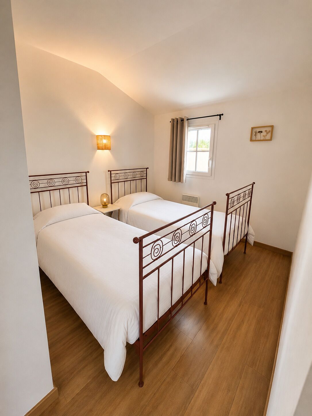 Une chambre avec deux lits, une fenêtre avec des rideaux, une lampe fixée au mur et un tableau.
