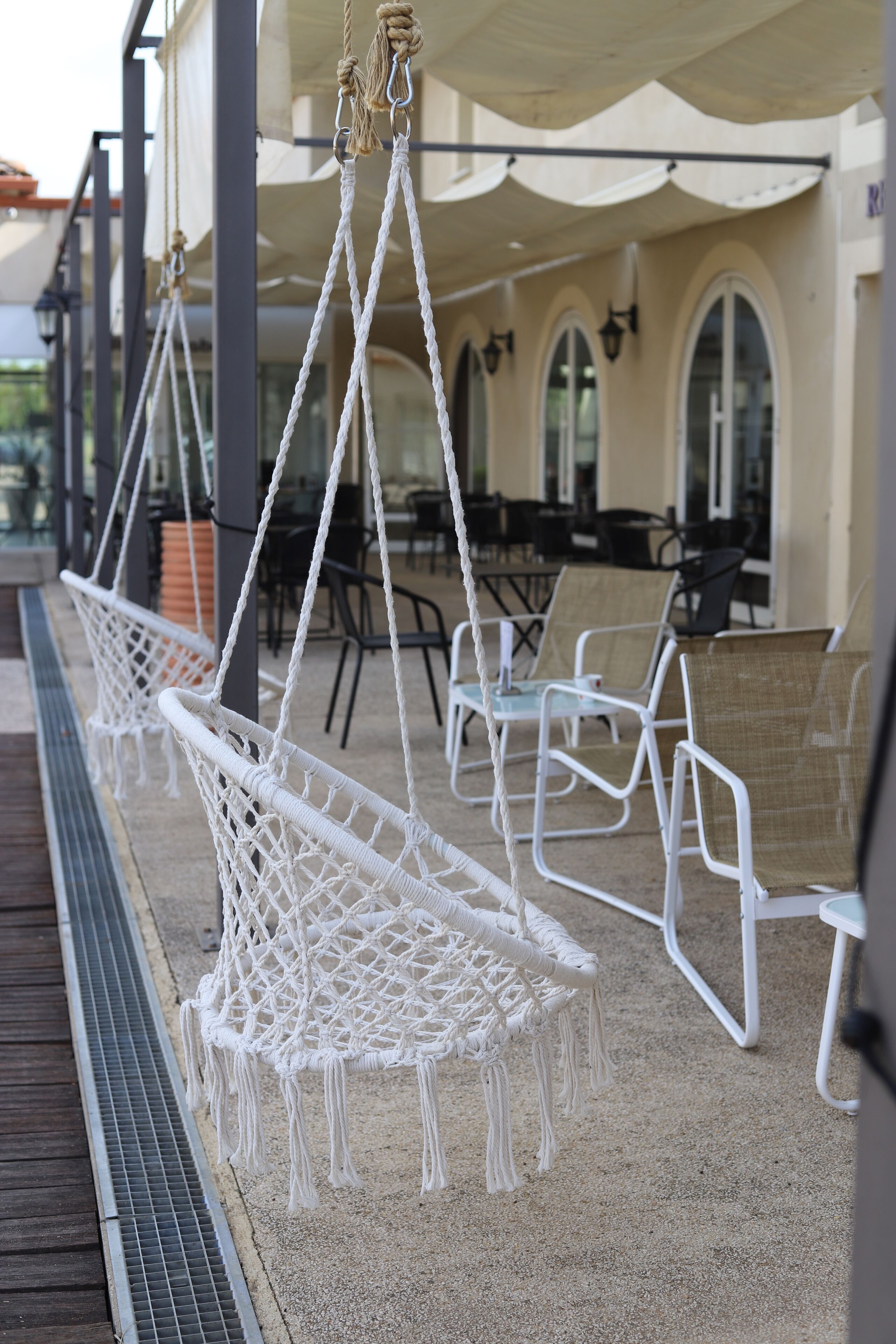 Espace extérieur aménagé avec des chaises et des cordes en macramé blanc, un patio couvert et, au fond, un bâtiment doté de fenêtres en arc de cercle.