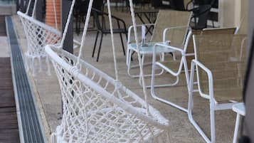 Espace extérieur aménagé avec des chaises et des cordes en macramé blanc, un patio couvert et, au fond, un bâtiment doté de fenêtres en arc de cercle.