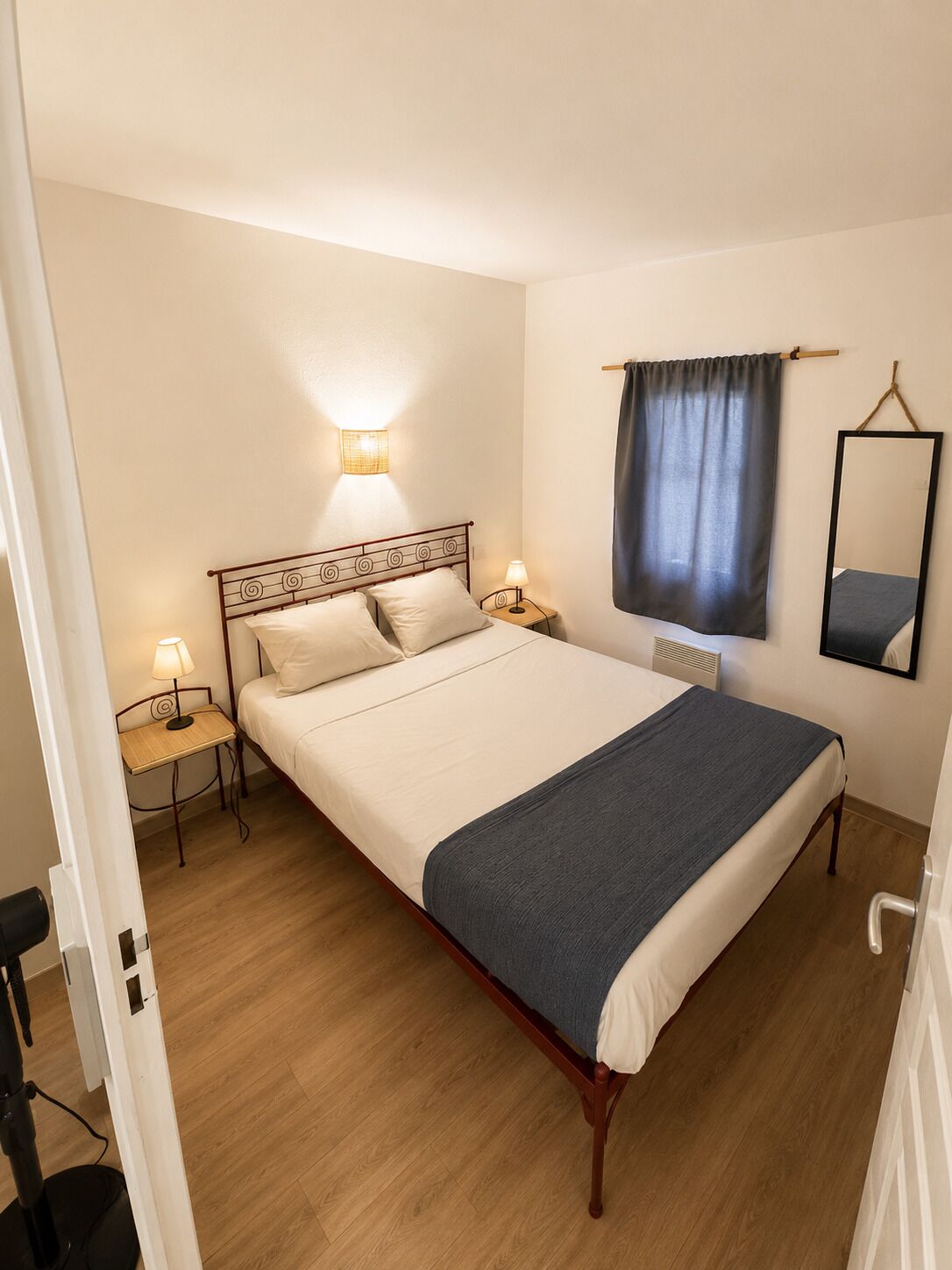 A single bed with a headboard, bedside tables with lamps, a wall-mounted mirror, and a window with a curtain.