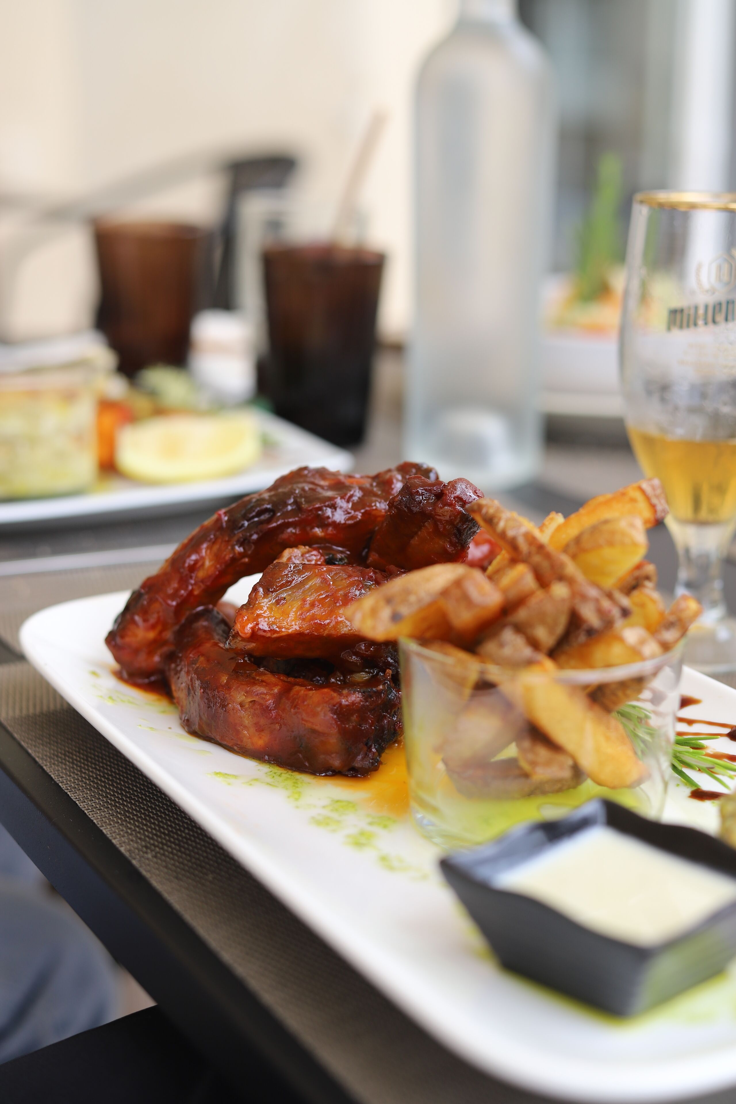A plate of ribs and fries with a small bowl of sauce, a glass of beer, and a bottle of water on a table.