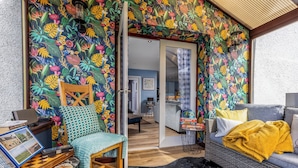 Una habitación con un vibrante papel tapiz tropical, un sofá de mimbre con una manta amarilla y una silla de madera con un cojín azul.
