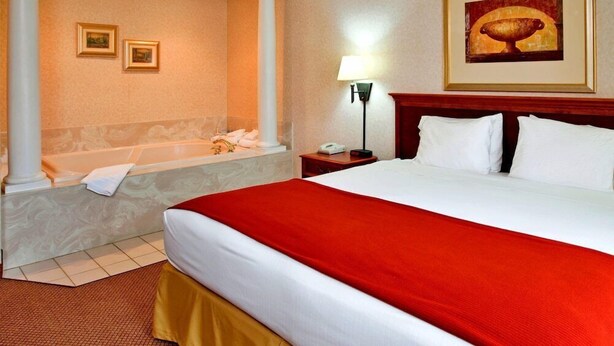 A hotel room with a large bed, a red bedspread, a bathtub, a nightstand with a lamp, and framed artwork on the wall. - Seasons Inn Suites Highland (Highland)