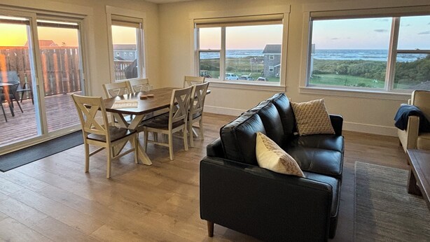 A spacious living area with a black leather sofa, wooden dining table, and chairs. Large windows offer views of the outdoors. - Ocean view cottages with direct beach access (Savage Harbor)