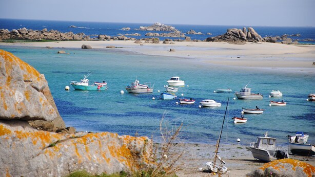 Un paysage côtier avec des bateaux ancrés près d’une plage de sable et de formations rocheuses.