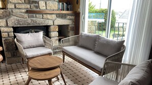 Un salon chaleureux avec une cheminée en pierre, des meubles en osier et une vue sur l’extérieur.