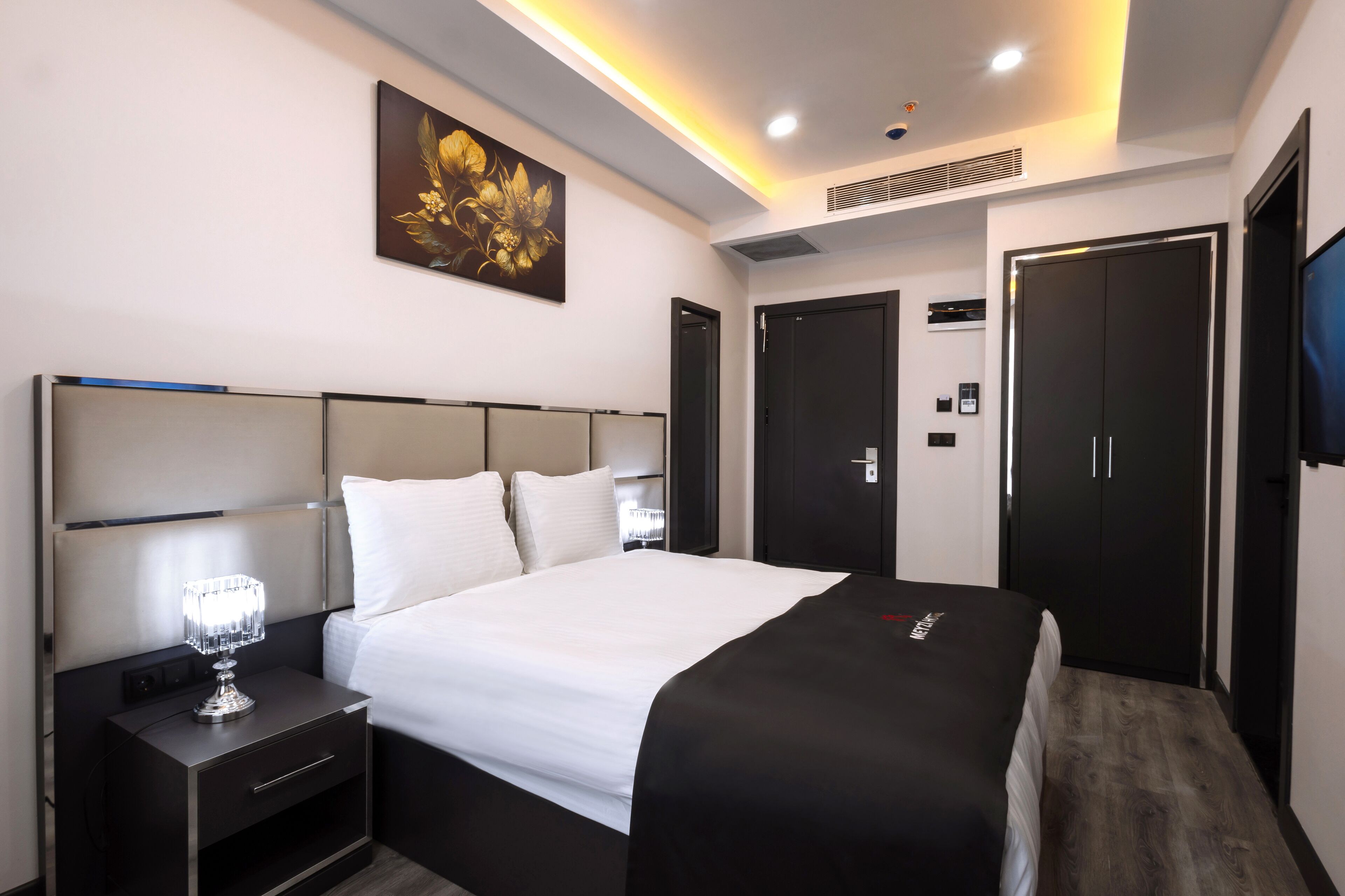 A modern hotel room with a large bed, a bedside table with a lamp, a wardrobe, and a painting on the wall.