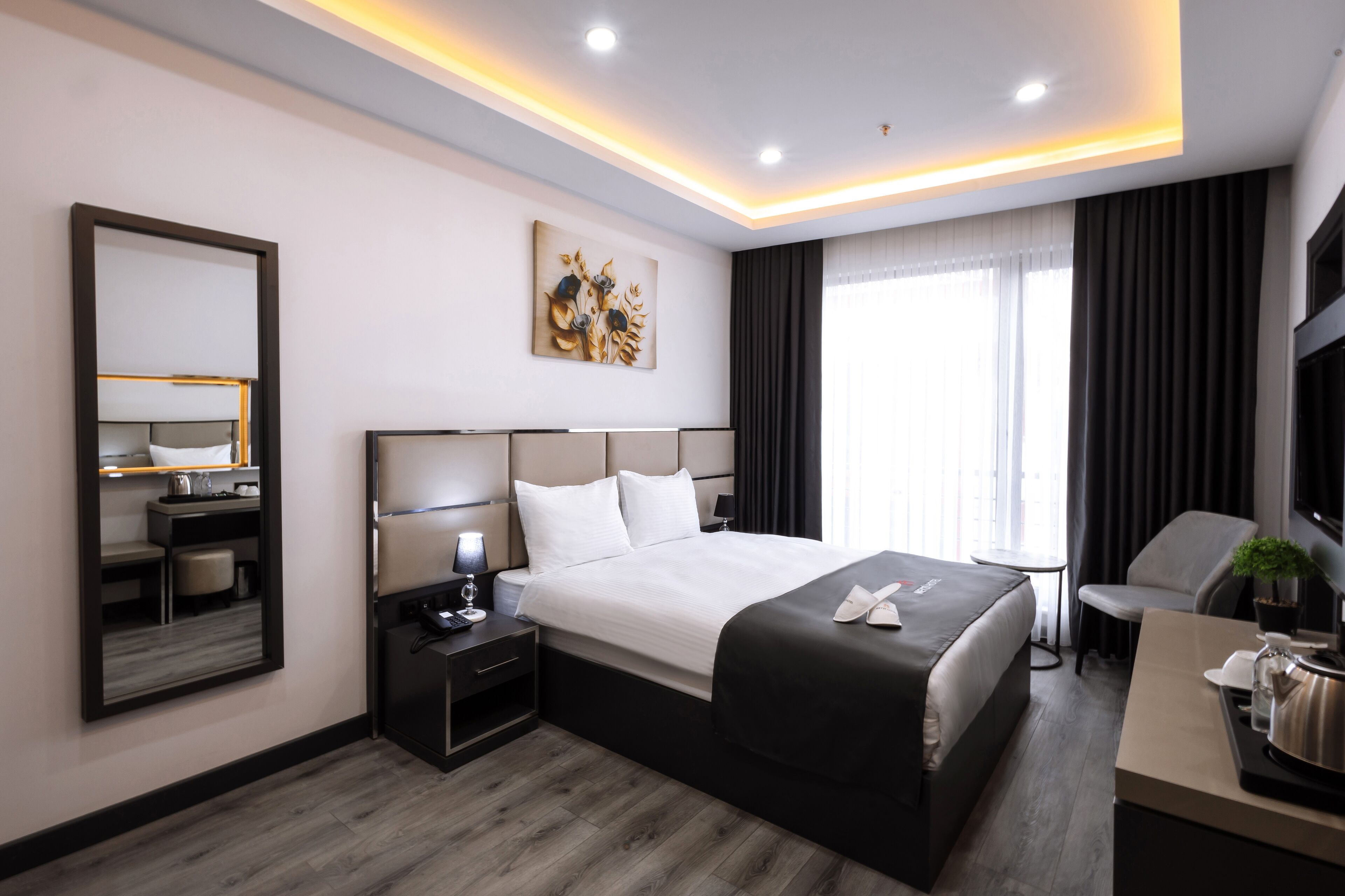 A modern hotel room with a large bed, bedside table, and a desk with a chair.