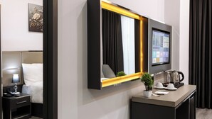 A modern hotel room with a large mirror, a flat-screen TV, a bedside table with a lamp, and a small plant. - Meyzi Hotel (Arnavutköy)