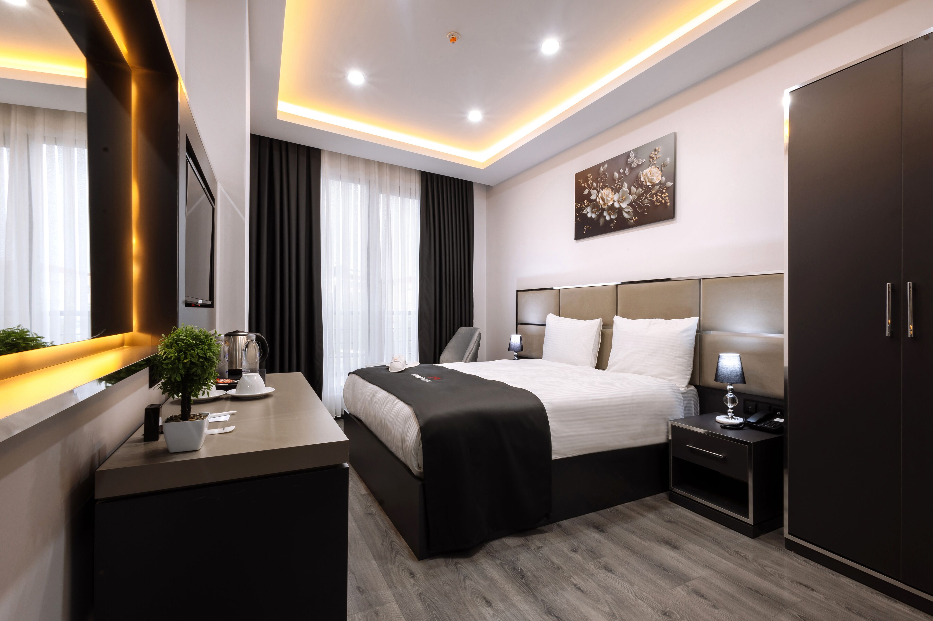 A modern hotel room with a large bed, bedside tables, a desk with a plant, a coffee maker, and a painting on the wall.