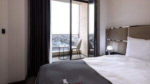 A hotel room with a large bed, a small table with a chair, a nightstand with a lamp, and a balcony with a view of the city. - Meyzi Hotel (Arnavutköy)