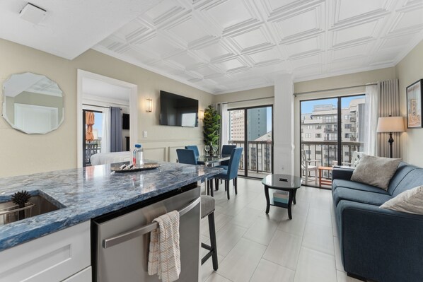 A modern apartment with a kitchen, dining area, and living room. It features a large window, a flat-screen TV, and a balcony with a view of the city.