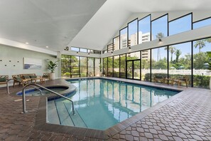 A modern indoor pool with clear glass walls, a brick floor, and wooden furniture.