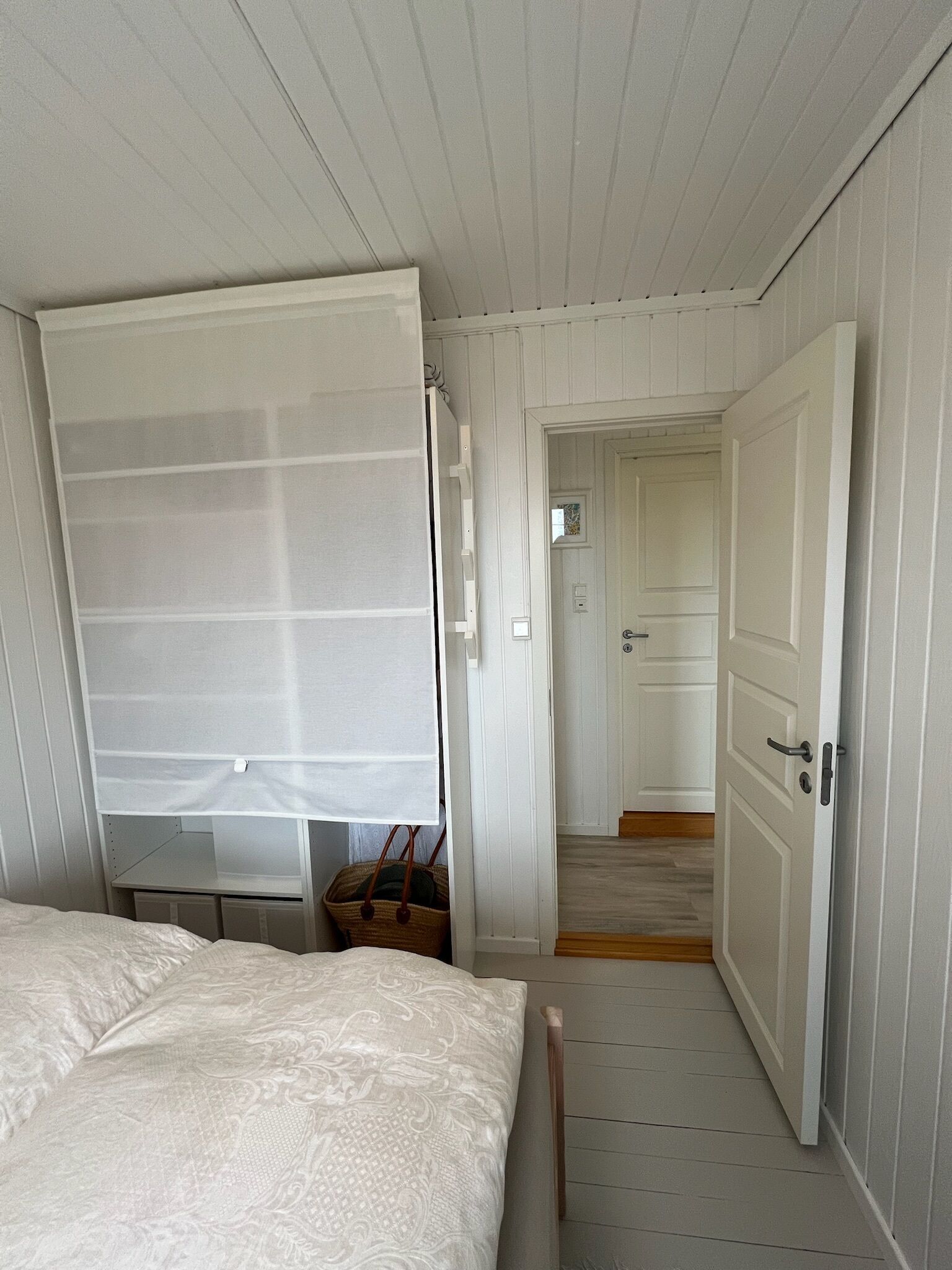 A bedroom with a bed, a window with a white blind, a wooden door, and a hallway leading to another room.