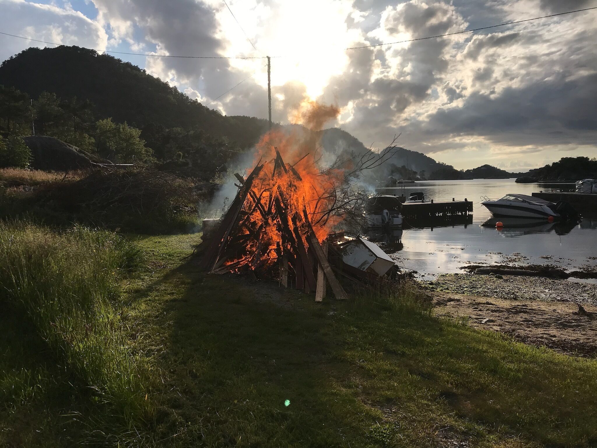 A campfire burning in a grassy area near a body of water with boats docked in the background.