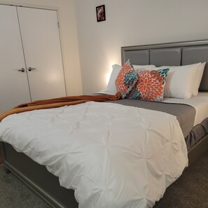 Cama bem arrumada com edredom branco e duas almofadas estampadas, cabeceira cinza e manta marrom.