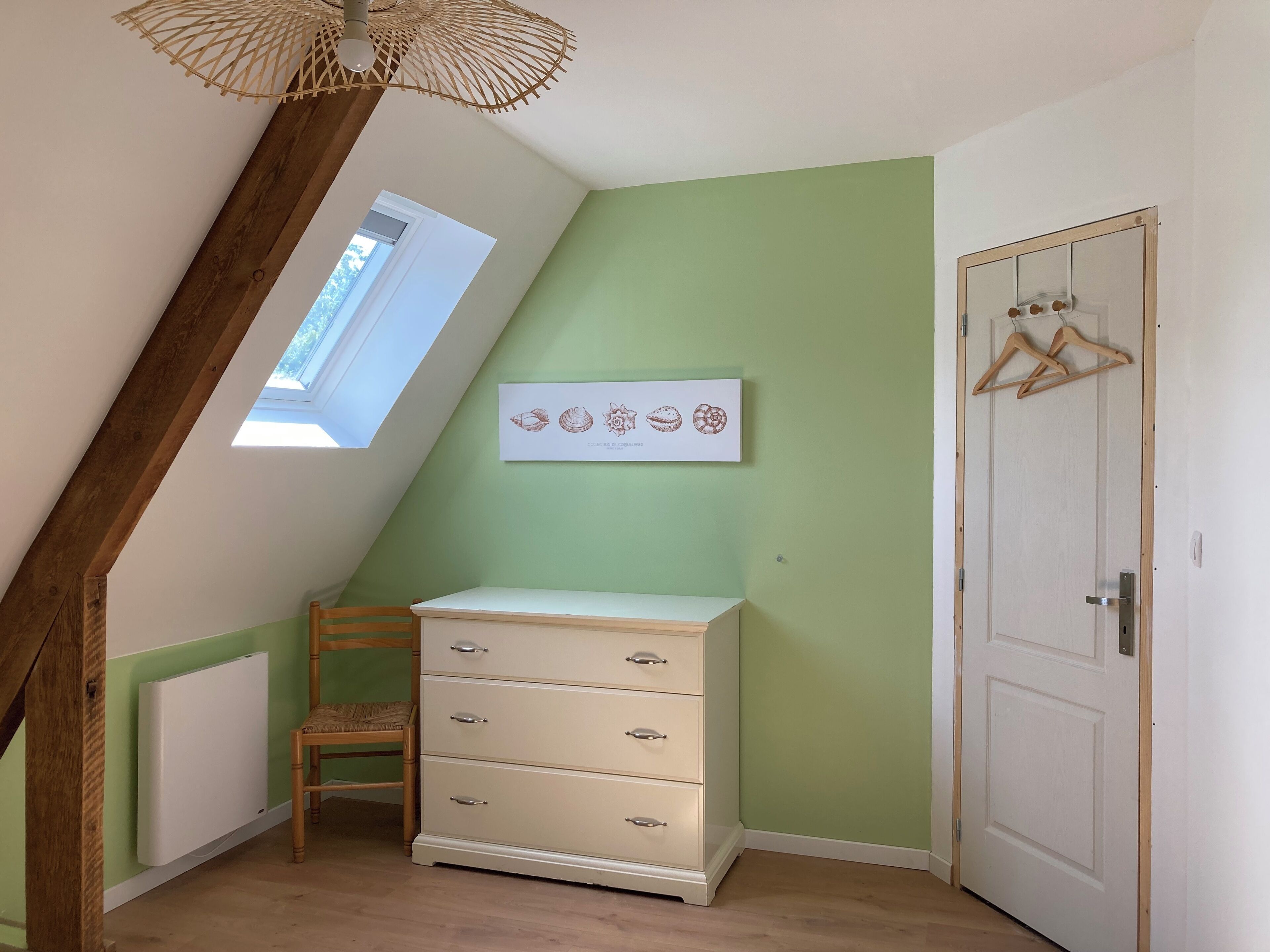 Ein Zimmer mit schrägem Dach, einem Holzstuhl, einer weißen Kommode, einer Tür und einem an der Wand befestigten Bild mit Muscheln.