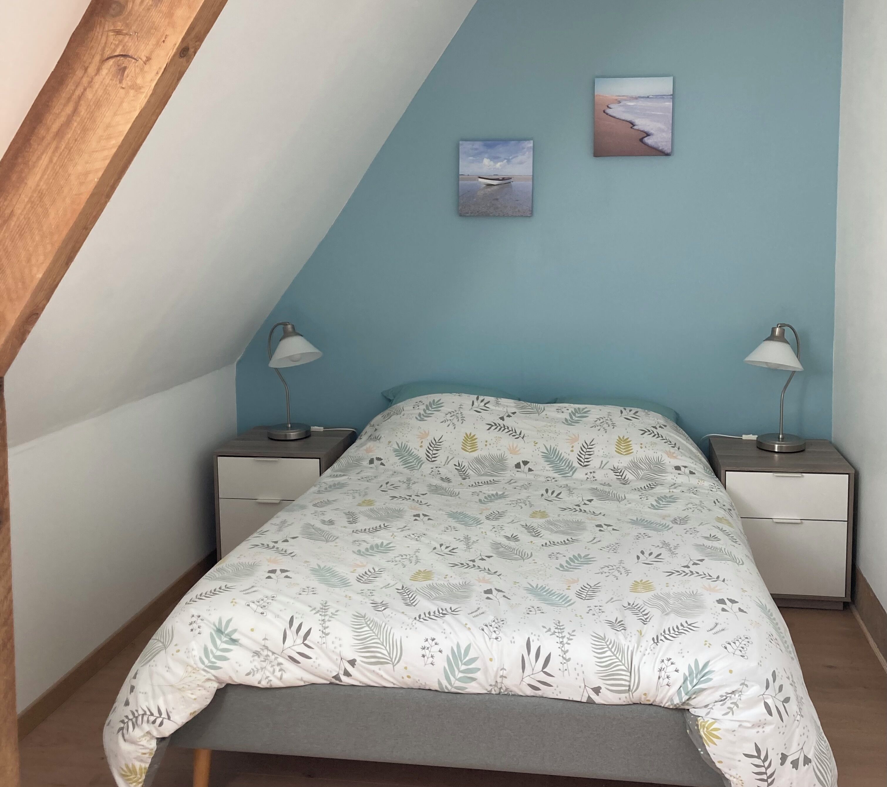 Een slaapkamer met een schuine kap, een bed met een sprei met bladmotief, twee nachtkastjes met lampen, en twee ingelijste foto's aan de muur.