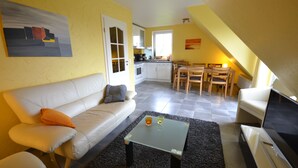 Ein Wohnzimmer mit einer Couch, einem Couchtisch und einem Fernseher. Im Hintergrund ist eine Küche mit einer Essecke zu sehen.