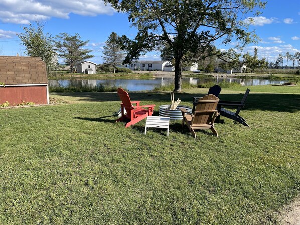 Un espacio al aire libre con césped, dos sillas Adirondack, un brasero de metal y un pequeño estanque al fondo.