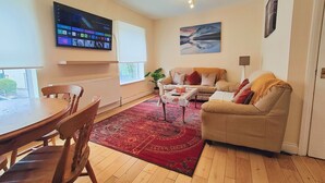 Ein Wohnzimmer mit einem beigefarbenen Sofa, einem roten Teppich mit Muster, einem Holzesstisch und einem an der Wand befestigten Fernseher.