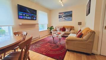 A living room with a beige sofa, a red patterned rug, a wooden dining table, and a TV mounted on the wall.