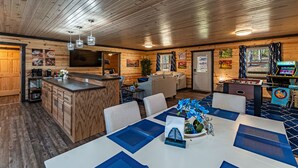 Un intérieur chaleureux, aménagé dans le style d'un chalet, avec un plafond en bois, un coin repas agrémenté de sets de table bleus, une cuisine avec un bar et un espace de détente équipé d'un baby-foot.