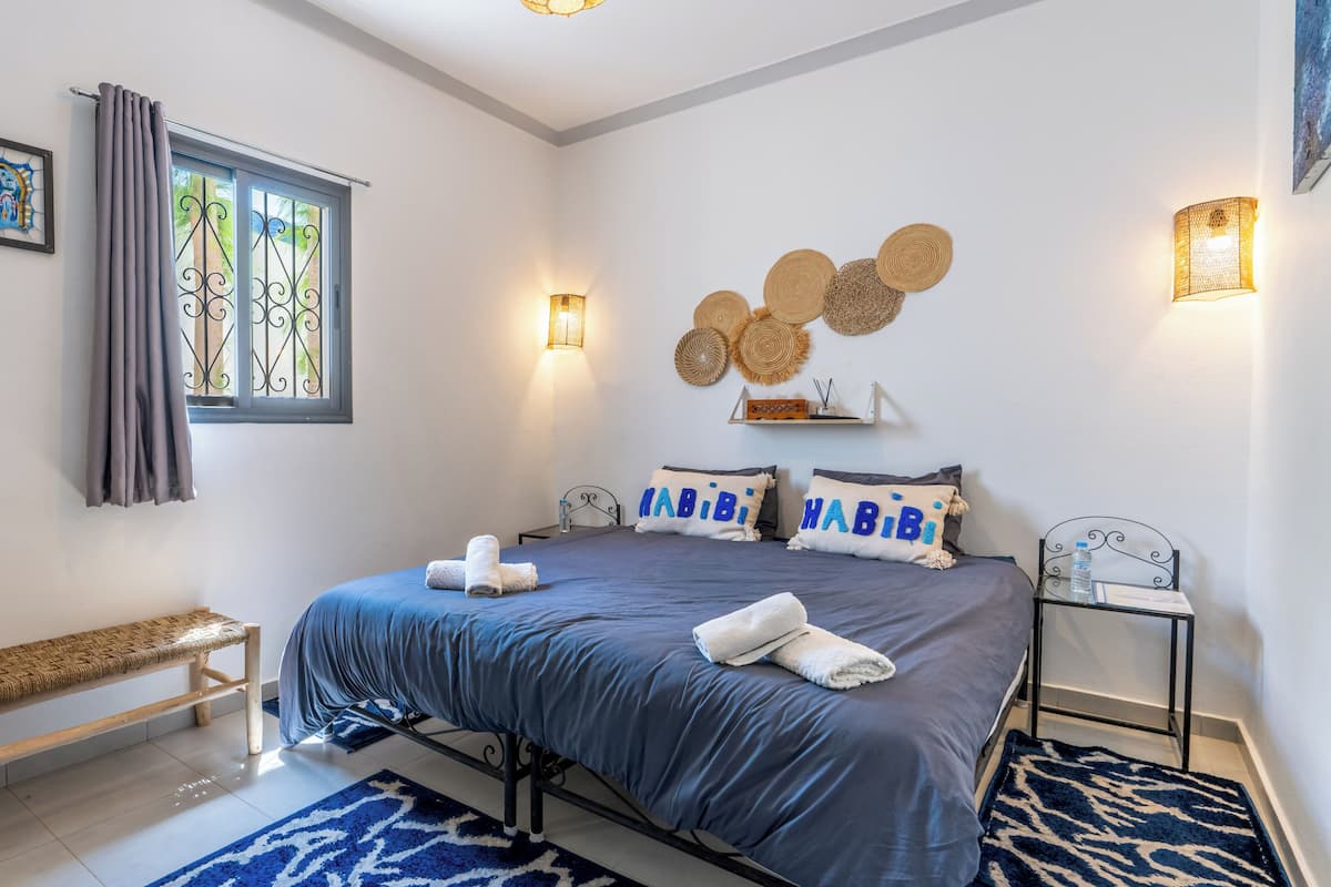 A bedroom with a bed, a bench, a wall-mounted shelf, and a window with curtains.