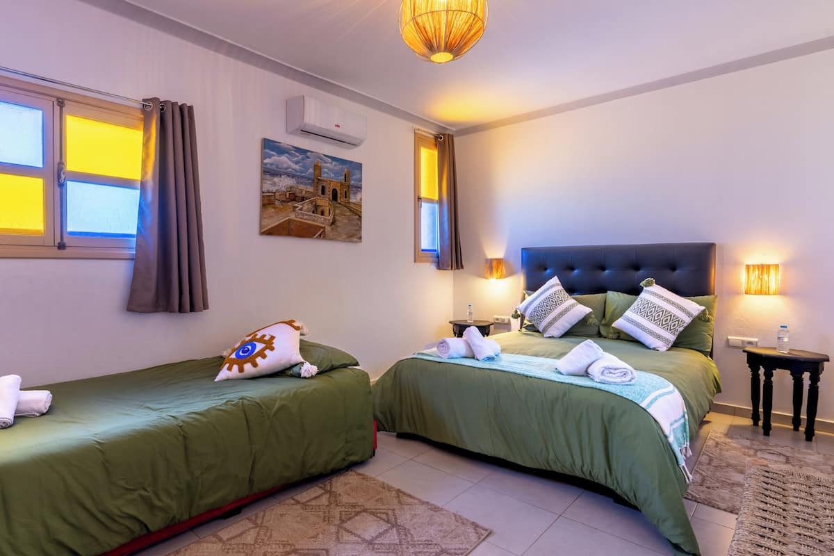 A hotel room with two beds, a window with curtains, a painting on the wall, an air conditioning unit, and a lamp on the nightstand.