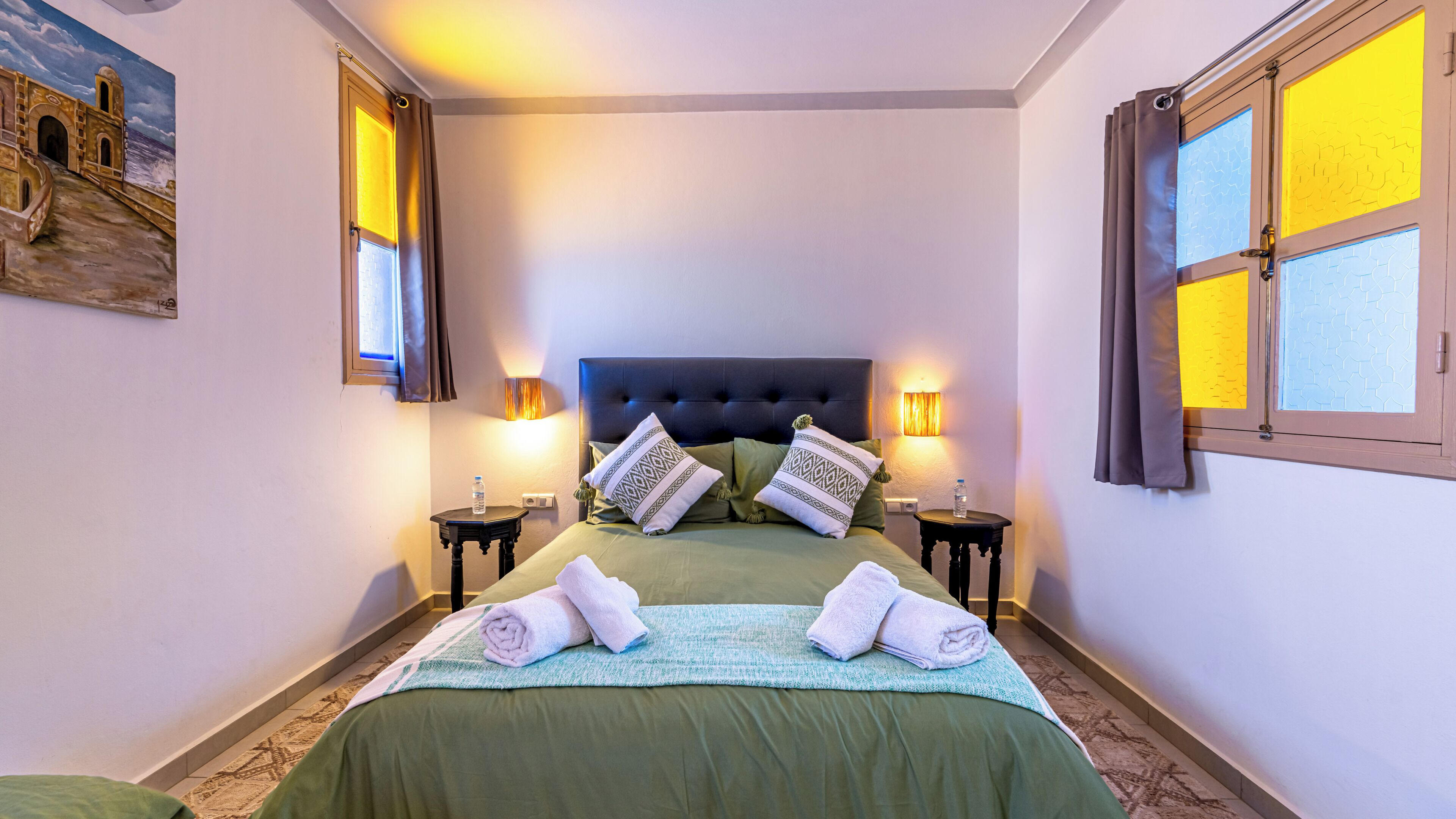 A hotel room with a large bed, two bedside tables, a headboard, and a painting on the wall.