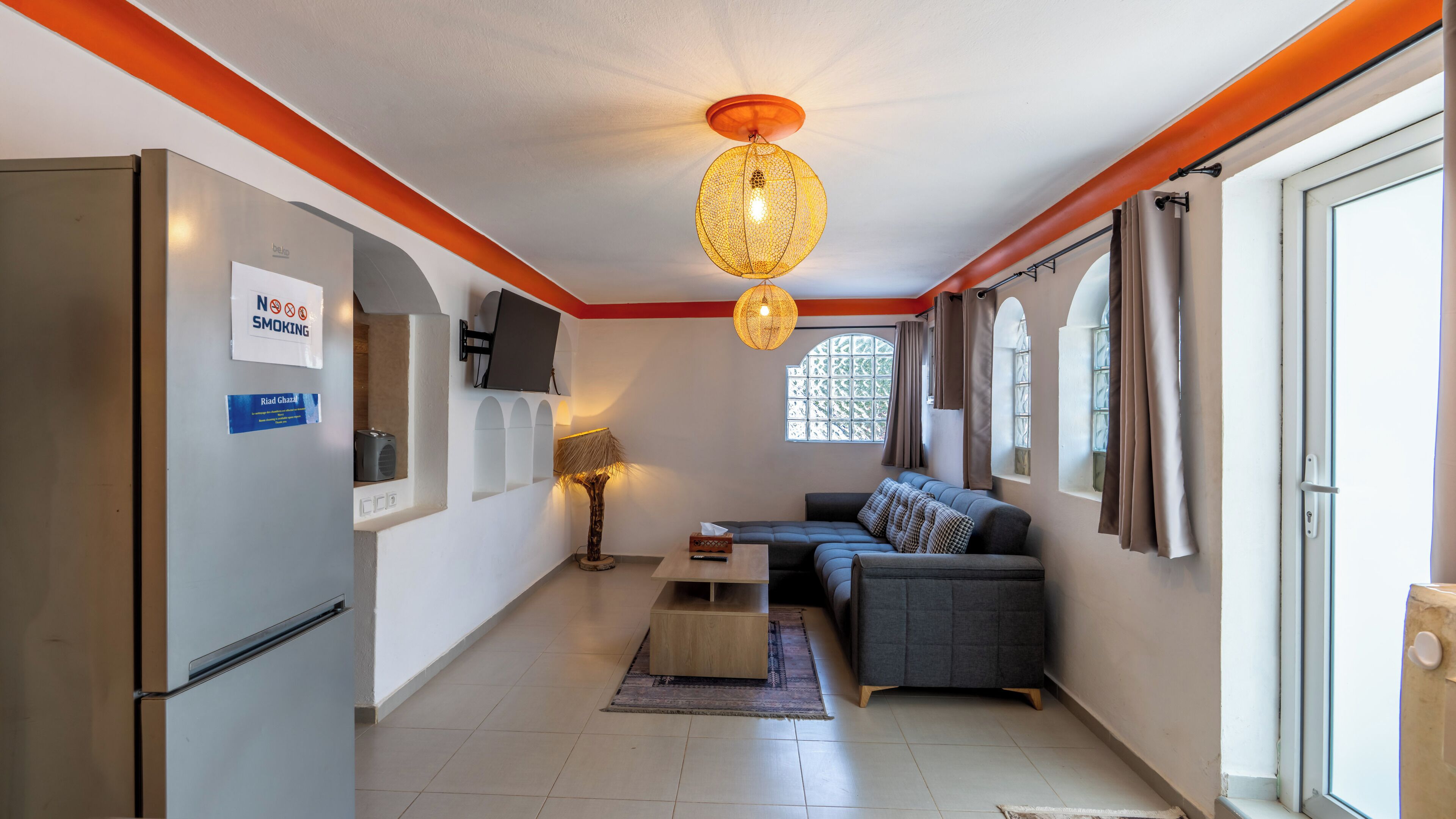 Um espaço compacto com sofá, televisão na parede, luminária no teto e janela com cortinas.